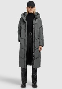 Lange grijze pufferjas met een oversized capuchon, rits- en knoopsluiting, geribbelde manchetten en zwarte accenten. Gedragen over een aansluitend zwart shirt en broek.