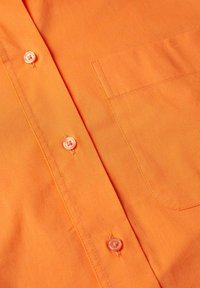 Devant d'une chemise en tissu orange avec des boutons blancs et une poche cousue sur le côté droit, montrant la texture et les détails des coutures.