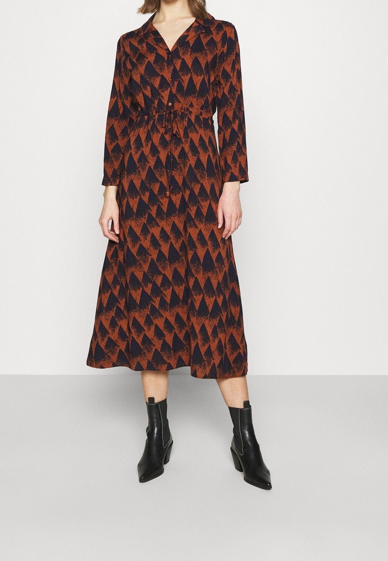 Robe à manches longues noire avec un motif géométrique orange, taille cintrée et jupe fluide. Associée à des bottines noires.
