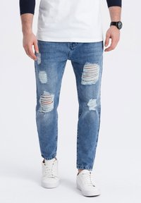 Vaqueros de mezclilla de color azul claro con múltiples áreas rasgadas, diseño ajustado y un acabado desgastado. Combinados con zapatillas blancas.