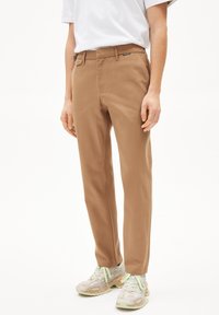 Pantalon beige avec une coupe droite, doté de passants de ceinture, de poches avant et d'une texture lisse, associé à des baskets blanches.