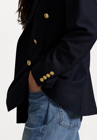 Sotilaallinen kaksirivinen bleiseri kultaisilla napilla, pehmeä kangas, rakenteellinen muotoilu ja hieman yli koko, yhdistettynä vaaleansinisiin farkkuihin.