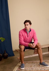 Roze kwart-rits pullover met een marineblauwe kraagdetail, gecombineerd met marineblauwe shorts en grijze schoenen, zittend op een houten bank. Gelaagde achtergrond.
