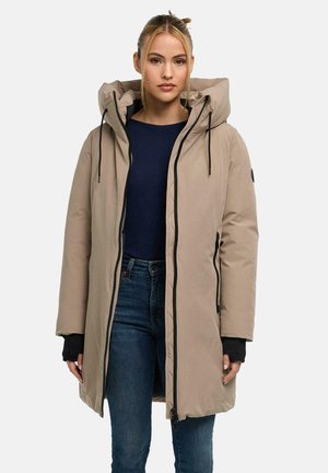 Donna che indossa un cappotto invernale beige con cappuccio sopra una maglia blu scuro e jeans blu, in piedi davanti a uno sfondo chiaro e uniforme.