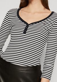 Mustavalkoraidallinen pitkähihainen paita, jossa on V-pääntie, kolmonen nappi kaula-aukossa ja pieni logo rinnassa.