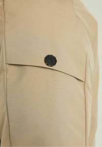 Beżowa tkanina o gładkiej fakturze z zapięciem na guzik. Projekt obejmuje klapową kieszeń z delikatnym połyskiem.