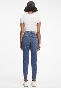 Jeans in denim blu a vita alta con due tasche posteriori, texture scolorita e un'etichetta in pelle marrone sulla vita, abbinati a una maglietta bianca.