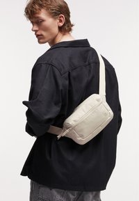 Borsa a tracolla beige con una texture liscia, design rettangolare e chiusura con zip, indossata sopra una maglietta nera da un uomo.