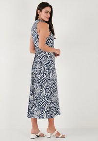 Sleeveless navy and white patterned midi dress with a fitted bodice, flared skirt, and a tie detail at the waist. Paired with white block-heel sandals.