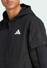 Black jacket with a hood, features a zip front, loose fit, and a pocket on the arm. Displayed logo is white. Smooth, textured fabric.