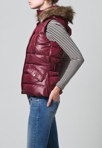 Gilet imbottito bordeaux con cappuccio in pelliccia staccabile, dotato di tasche frontali, vita aderente e finitura lucida. Indossato sopra una maglia a maniche lunghe a righe.