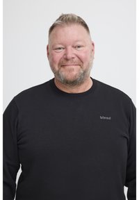 Uomo di mezza età sorridente con capelli corti biondi e barba, indossa un felpa nera con il logo "blend" su uno sfondo bianco.