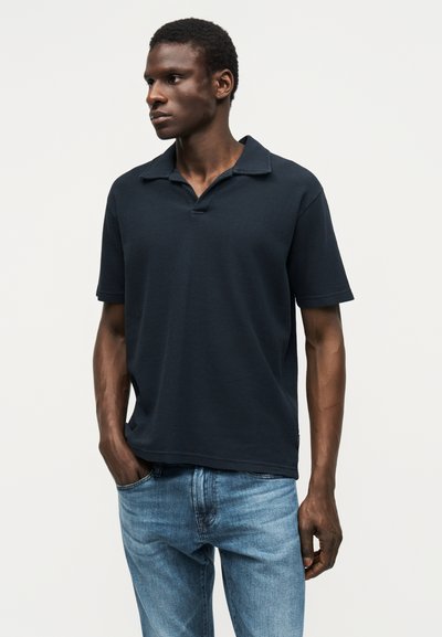 Homme portant un polo noir à manches courtes et un jean bleu clair, debout avec une main dans la poche, regardant vers la gauche.