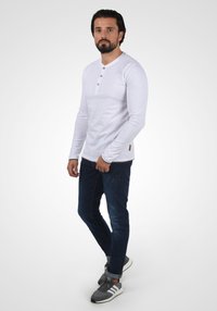 Hvit langermet henley-skjorte med tre brune knapper, paret med mørkeblå jeans og grå sneakers med hvite såler og oransje detaljer.