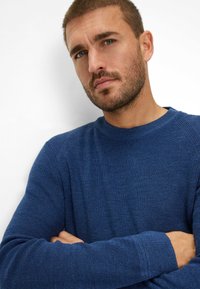Blauwe gebreide trui met een gestructureerde afwerking, ronde halslijn en geribbelde manchetten. Model heeft de armen gekruist en poseert tegen een witte achtergrond.