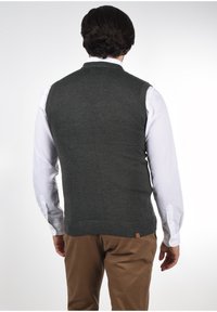 Gilet gris en tricot avec col rond, porté par-dessus une chemise blanche. Présente un ourlet côtelé et une étiquette en bas.