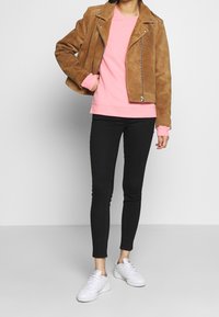 Person, der eine braune Wildlederjacke über einem pinken Sweatshirt, schwarzen Skinny-Jeans und weißen Sneakers trägt, steht vor einem schlichten Hintergrund.