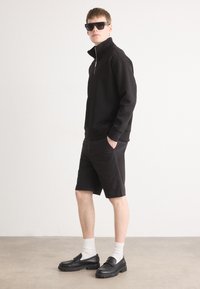 Sweat à zip noir en polaire associé à un short noir. Le modèle porte des lunettes de soleil noires et des mocassins noirs avec des chaussettes blanches sur un fond neutre.