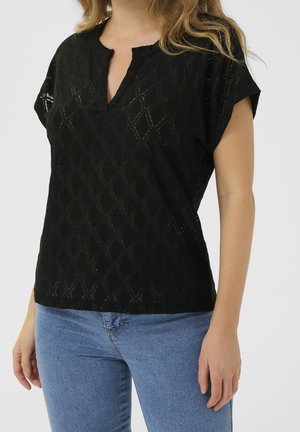 Kvinde iført en sort kortærmet bluse med diamantmønstre og blå jeans mod en ensfarvet baggrund.