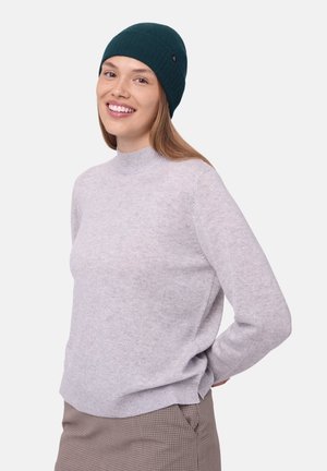 Harmaa neulepusero korkealla kauluksella; mukana tummanvihreä pipo. Malli hymyilee, antaen esiin pehmeän tekstuurin ja rennon istuvuuden. Ruudullinen hame näkyvissä.