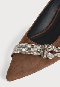Chaussure plate en daim marron pointue avec une bride scintillante en strass nouée en nœud sur le dessus.