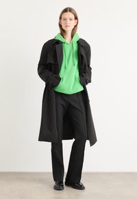 Palton negru peste un hanorac cu glugă verde, asortat cu pantaloni negri ajustați și pantofi. Texturi netede cu linii clare.