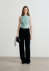 Γυναίκα που φοράει αμάνικο ανοιχτοπράσινο τοπ και μαύρο παντελόνι με φαρδιά πόδια, κρατώντας μια μικρή μαύρη τσάντα, πάνω σε απλό λευκό φόντο.