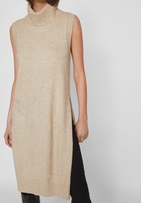 Beige ärmlös turtleneck-tunika med ribbad krage, sidoklipp och mjuk textur, tillsammans med svarta byxor.