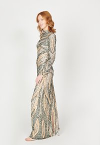 Robe longue et ajustée avec manches longues, ornée de motifs de sequins dorés et verts évoquant des feuilles, et avec une fente sur le côté pour faciliter les mouvements.