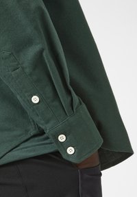 Donkergroene shirt van een gestructureerde stof. Lange mouwen met witte knopen bij de manchetten en zijsplitdetails.