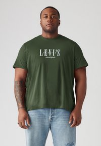 Olive spalvos medvilnės marškinėliai su balta grafika ir "LEVI'S 1873" tekstu, trumpomis rankovėmis ir laisvu siluetu. Dėvimi su šviesiai mėlynomis nusidėvėjusiomis džinsais.