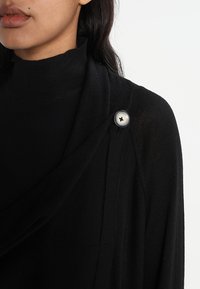 Pull noir avec un design drapé, doté d'un grand bouton métallique sur l'épaule et d'un tissu tricoté doux et texturé.