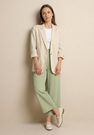 Vrouw staat tegen een beige muur, draagt een crème blazer, witte top, lichtgroene wijde broek en witte loafers met gouden kettingdetail.