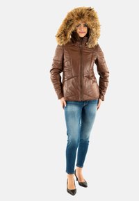 Chaqueta de cuero marrón con capucha de piel sintética, que presenta un cierre frontal con cremallera, diseño acolchado y bolsillos. Se lleva sobre vaqueros azules y tacones negros.