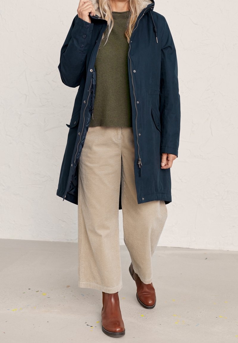 Granatowy wodoodporny płaszcz z kapturem wyściełanym futrem, zamkiem błyskawicznym i regulowanymi sznureczkami, noszony na oliwkowozielonym swetrze i beżowych spodniach sztruksowych.