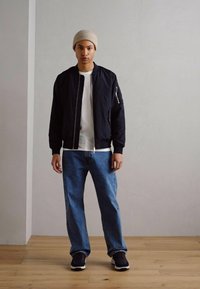 Giacca bomber nera sopra una maglietta bianca, abbinata a jeans azzurri chiari e sneakers nere. Un berretto lavorato a maglia beige completa l'outfit.