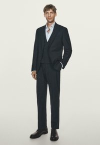 Dunkelblauer Dreiteiler mit einem taillierten Blazer, passenden Hosen und einem hellblauen Button-Down-Hemd. Schwarze Lederschuhe vervollständigen den Look.