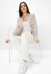 Gestreiftes Button-up-Hemd in Beige und Weiß, kombiniert mit einem weißen Tanktop und weißen Schlaghosen. Weiße Sneaker rundet das Outfit ab.