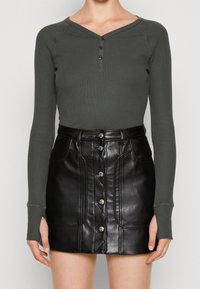 Olivgrünes, geripptes Langarm-Oberteil mit V-Ausschnitt und drei dunklen Knöpfen, kombiniert mit einem schwarzen, faux-ledernen Miniskirt mit Knopfleiste und Nähten.