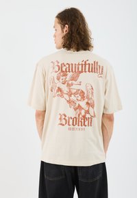Beige bavlnené tričko s podrobným rust-red potlačou cherubínov a textom "Beautifully Broken" na chrbte, s krátkymi rukávmi a voľným strihom.
