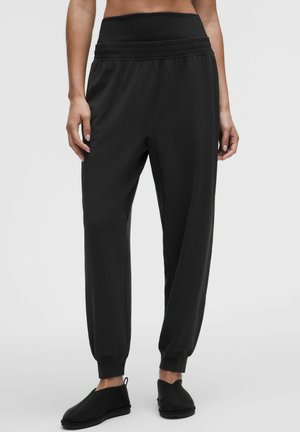 Vrouw die zwarte joggerbroek met hoge taille en elastische boorden draagt, gecombineerd met zwarte instapschoenen, staand tegen een effen achtergrond.