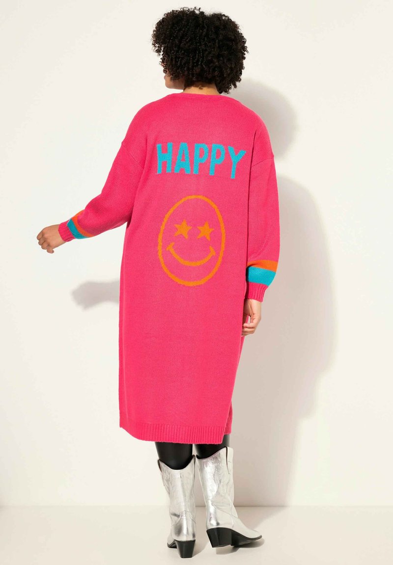 Grosse cardigan tricoté rose avec le texte "HAPPY" et un design de smiley au dos. Les poignets présentent des rayures multicolores. Porté avec des bottes argentées.