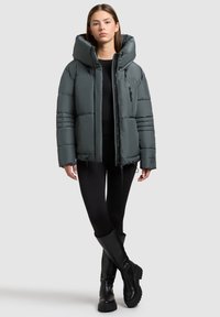 Ung kvinde iført en grå polstret jakke, sorte leggings og sorte knæhøje støvler, stående med krydsede ben foran en ensfarvet baggrund.