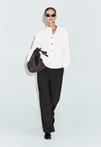 Witte langwerpige shirt met knopenrij, gecombineerd met zwarte wijde broek en een donkerbruine tas. Zonnebril maakt de look compleet.