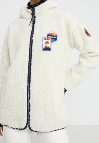 Vit fleece zip-up jacka med huva, marinblå detaljer och broderade lappar med landskap och varumärkeslogotyp på bröstet.