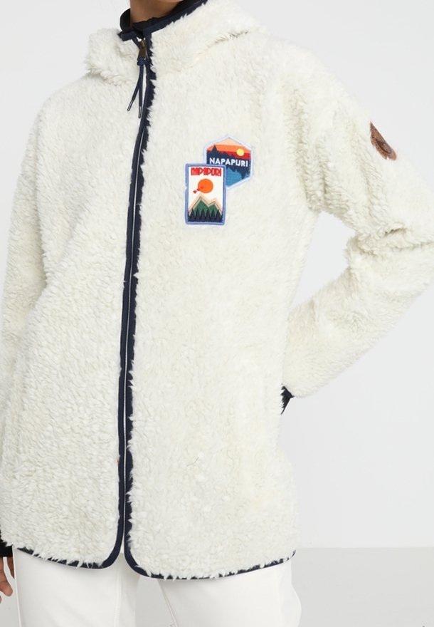 Vit fleece zip-up jacka med huva, marinblå detaljer och broderade lappar med landskap och varumärkeslogotyp på bröstet.