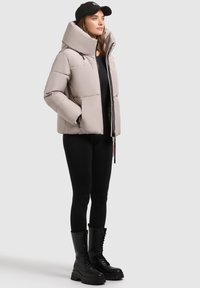 Gepolsterte beige Jacke mit hohem Kragen und Reißverschluss vorne, kombiniert mit schwarzen Leggings und knielangen Schnürstiefeln. Glatte Textur, lässiges Design.