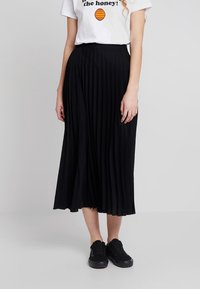 Svart plisserad midikjol av lättviktsmaterial, med hög midja och slät yta, kombinerad med en vit grafisk t-shirt och svarta sneakers.