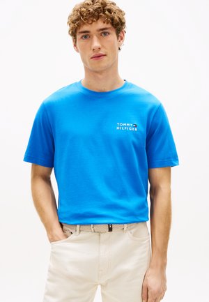 Jeune homme aux cheveux bouclés portant un t-shirt bleu vif Tommy Hilfiger et un pantalon beige, debout avec une main dans la poche.