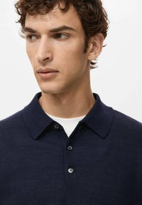 Młody mężczyzna z kręconymi brązowymi włosami, ubrany w granatowy sweter na guziki na białą koszulę, patrzący w lewo na białym tle.